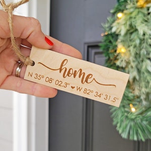 May include: A wooden home decor sign with the word "home" in cursive script and coordinates engraved below. The coordinates are N 35° 08' 02.3" W 82° 34' 31.5".