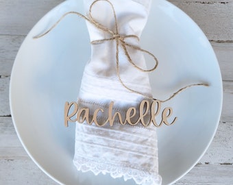 May include: A white napkin with a lace trim is tied with twine and a wooden name card that reads "Rachelle". The napkin is on a white plate.