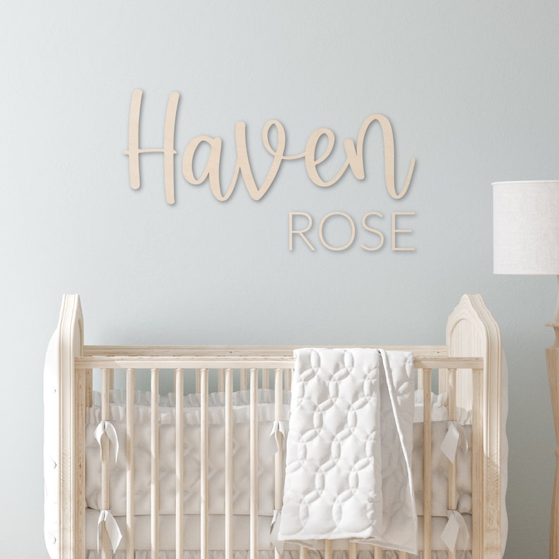 May include: A white wooden sign with the name "Haven Rose" written in cursive script. The sign is hanging on a light blue wall above a white cot.