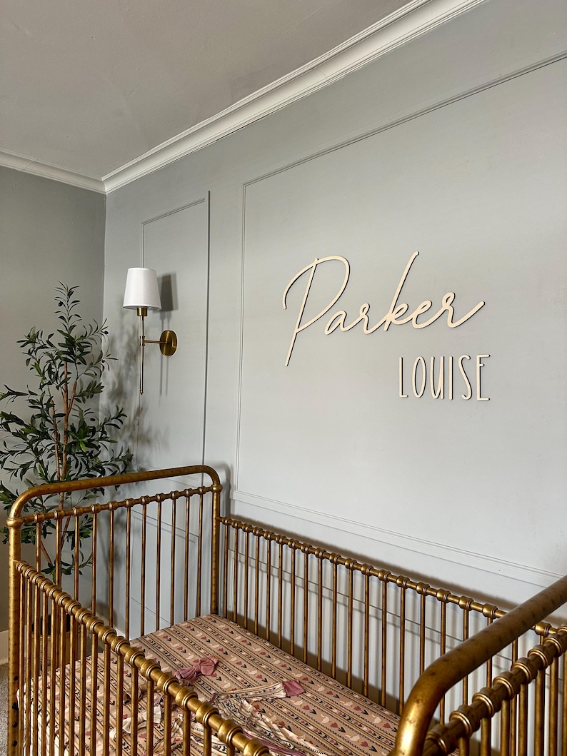 Puede incluir: Una escena de guarder&iacute;a con una cuna dorada y un colch&oacute;n estampado. La pared gris claro muestra el nombre "Parker Louise" en escritura de madera. Tambi&eacute;n se ven un aplique de pared y una planta en maceta.