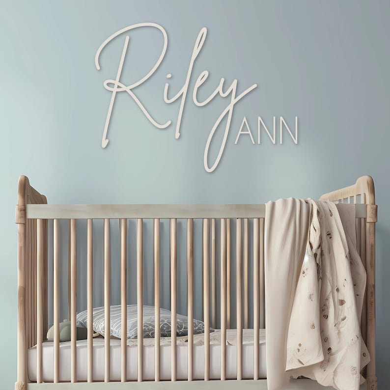 Puede incluir: Escena de guarder&iacute;a con una cuna de madera contra una pared azul claro. El nombre "Riley Ann" se muestra en una fuente cursiva encima de la cuna. La cuna tiene un colch&oacute;n, una almohada y una manta sobre el lateral.