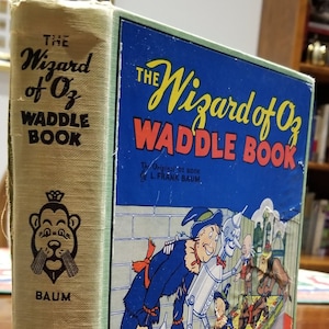 May include: A vintage hardcover book titled "The Wizard of Oz Waddle Book" by L. Frank Baum. The cover features a colorful illustration of the Scarecrow, Tin Man, and Dorothy walking down the Yellow Brick Road.