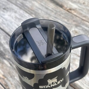 Op de afbeelding: Een zwarte en zilveren Stanley-reistumbler met een zwart vleermuispatroon en een zwart deksel met een grijze schuif. De tumbler heeft een zwarte handgreep en een roestvrijstalen rietje.