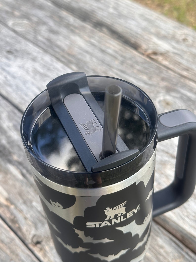 Op de afbeelding: Een zwarte en zilveren Stanley-reistumbler met een zwart vleermuispatroon en een zwart deksel met een grijze schuif. De tumbler heeft een zwarte handgreep en een roestvrijstalen rietje.