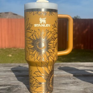 K&ouml;nnte beinhalten: Eine goldene Edelstahl-Reisebecher mit einem schwarzen Sonnenblumenmuster und dem Wort "STANLEY" auf der Seite. Der Becher hat einen Deckel mit einem Strohhalm und einem Griff.