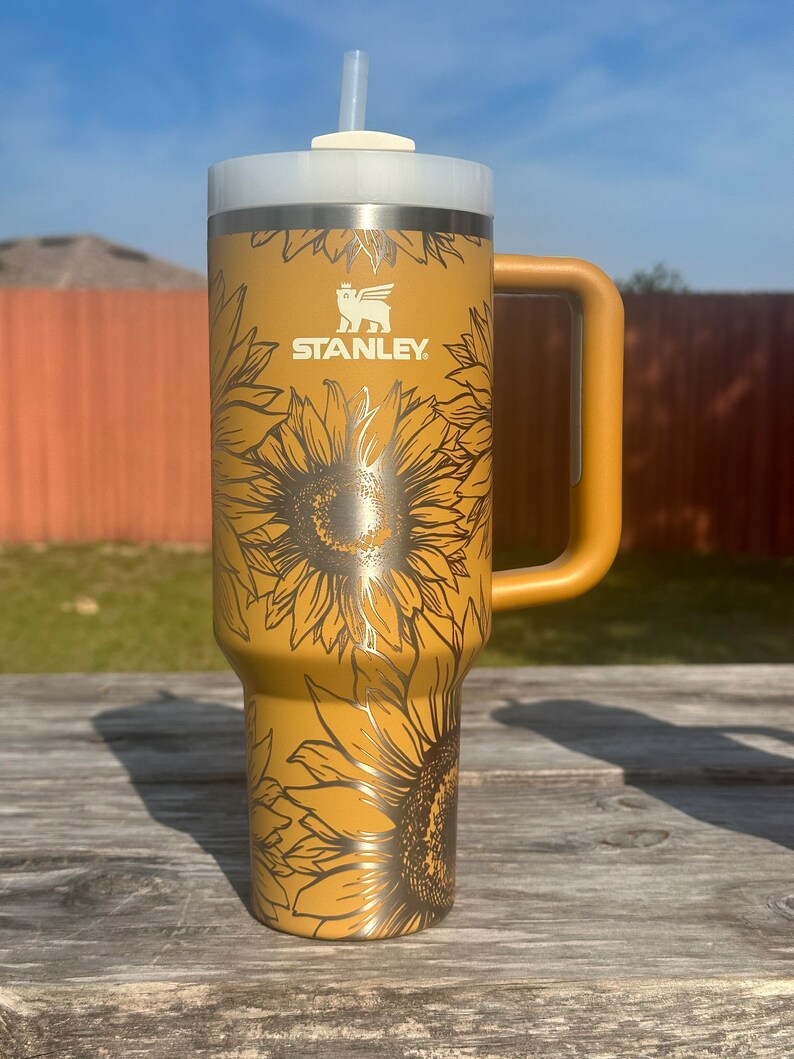 K&ouml;nnte beinhalten: Eine goldene Edelstahl-Reisebecher mit einem schwarzen Sonnenblumenmuster und dem Wort "STANLEY" auf der Seite. Der Becher hat einen Deckel mit einem Strohhalm und einem Griff.