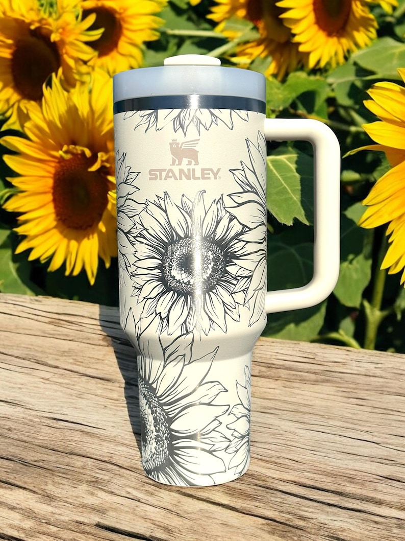 K&ouml;nnte beinhalten: Wei&szlig;e Edelstahl-Reisebecher mit einem schwarz-wei&szlig;en Sonnenblumenmuster. Der Becher hat einen schwarzen Deckel und einen schwarzen Griff. Der Becher tr&auml;gt das Stanley-Logo auf der Vorderseite.