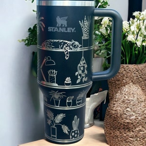 May include: A black Stanley travel mug with a silver handle and a silver cat and plants design. The mug has the Stanley logo and the text "Stanley" on the side.