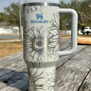 K&ouml;nnte beinhalten: Ein wei&szlig;er Edelstahlbecher mit einem schwarz-wei&szlig;en Sonnenblumenmuster. Der Becher hat einen Griff und einen Strohhalm. Der Becher tr&auml;gt das Stanley-Logo.