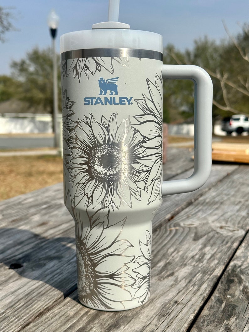 K&ouml;nnte beinhalten: Ein wei&szlig;er Edelstahlbecher mit einem schwarz-wei&szlig;en Sonnenblumenmuster. Der Becher hat einen Griff und einen Strohhalm. Der Becher tr&auml;gt das Stanley-Logo.