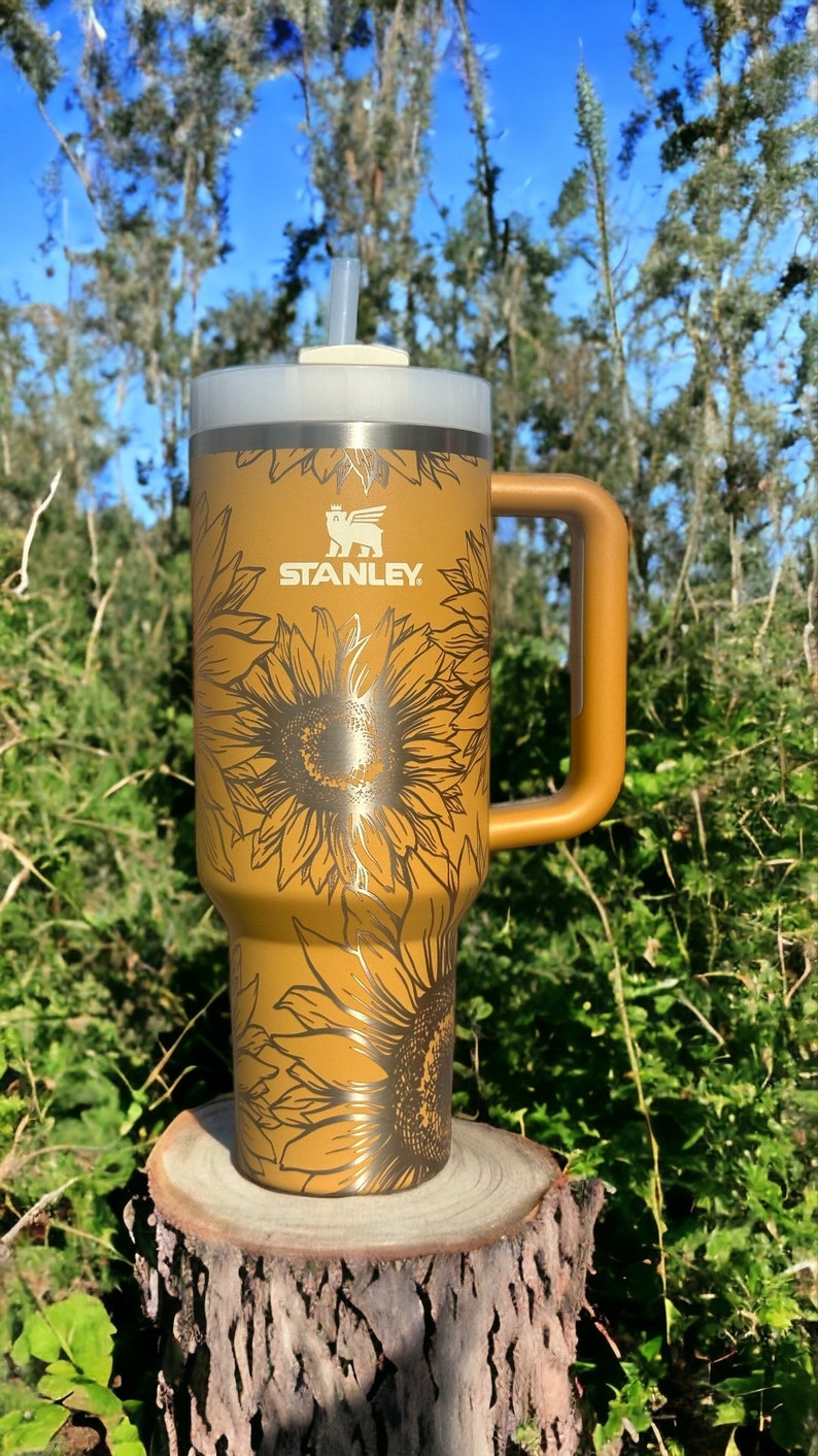 K&ouml;nnte beinhalten: Ein goldfarbener Edelstahlbecher mit einem Sonnenblumenmuster und dem aufgedruckten Wort "STANLEY". Der Becher hat einen Deckel mit einem Strohhalmloch und einen Griff.