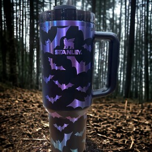 Op de afbeelding: Een zwarte reistumbler van roestvrij staal met een vleermuispatroon en een zwarte handgreep. De tumbler heeft een doorzichtig deksel met een rietje en het Stanley-logo is aan de zijkant gedrukt.