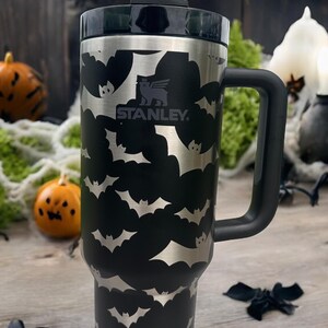 Op de afbeelding: Een zwarte roestvrijstalen reistumbler met een zwarte handgreep en een zwarte deksel. De tumbler heeft een vleermuispatroon en het Stanley-logo. De tumbler wordt geleverd met een rietje.