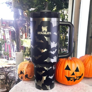 Op de afbeelding: Zwarte reistumbler van roestvrij staal met een vleermuispatroon en het Stanley-logo. De tumbler heeft een zwarte handgreep en een zwarte deksel met een rietje.