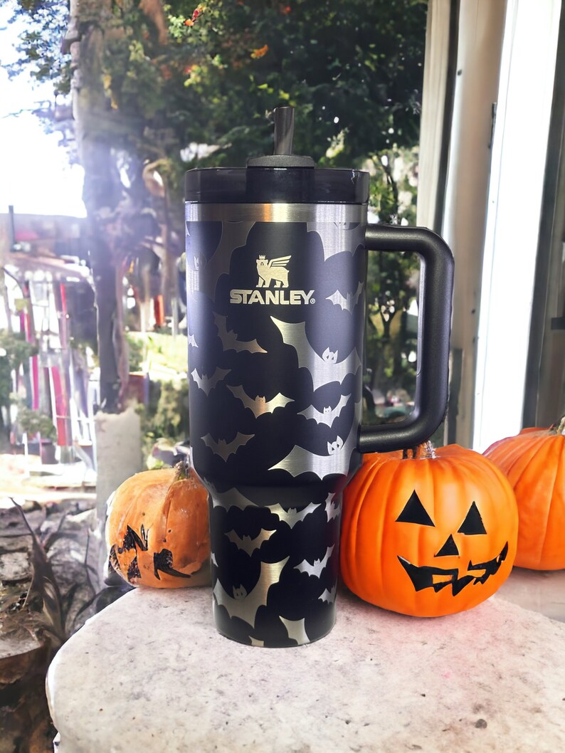 Op de afbeelding: Zwarte reistumbler van roestvrij staal met een vleermuispatroon en het Stanley-logo. De tumbler heeft een zwarte handgreep en een zwarte deksel met een rietje.