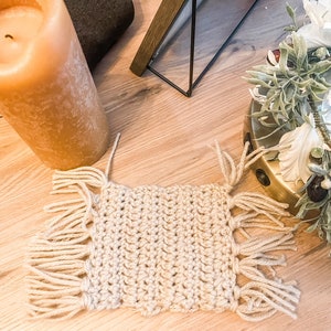 May include: A beige crocheted coaster with fringe around the edges. The coaster is on a wooden surface.