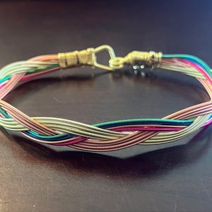 May include: A braided bracelet made from colorful metal wires. The bracelet features a gold-colored clasp and is composed of various shades, including pink, green, blue, and gold. The design is intricate and eye-catching, suitable for everyday wear.