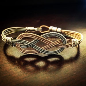 May include: A handcrafted bracelet featuring an infinity knot design. The bracelet is made from silver and gold-colored wire, with a woven pattern forming the knot. The clasp and connecting elements are silver. The bracelet is displayed on a dark surface.
