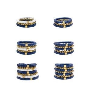 May include: Set of six stacked bracelets with blue and white beads and gold accents. The bracelets are arranged in three pairs, with each pair featuring a different combination of colors and beads.