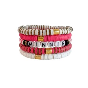 May include: A set of four bracelets with white, pink, and gold beads. The middle bracelet has white beads with the word "MINNIE" spelled out in black.