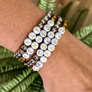May include: Four white beaded bracelets with gold lettering spelling out the names "Katelyn", "Cody", "Cam", and "Campbell". The bracelets are stacked on a person's wrist.