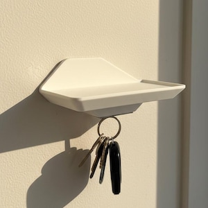 May include: A white, geometric key holder mounted on a wall. The holder has a small shelf and a metal ring holding several keys. The shadow of the holder is cast on the wall.
