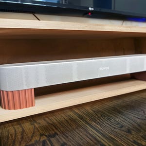 May include: A gray soundbar with the brand name "SONOS" in white lettering. The soundbar is supported by two coral-colored, ribbed stands. The soundbar is placed on a wooden shelf, with a TV visible in the background.