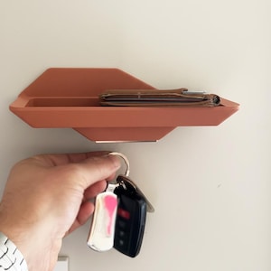 May include: A terracotta-colored wall-mounted shelf with a geometric design. A brown leather wallet rests on the shelf. A hand holds a set of keys with a silver fob and a black car key.