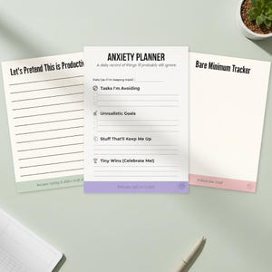 May include: Three white notepads with humorous text. The center notepad is labeled "Anxiety Planner" with prompts for tracking tasks, goals, and wins. The other notepads are labeled "Let's Pretend This is Productive" and "Bare Minimum Tracker".