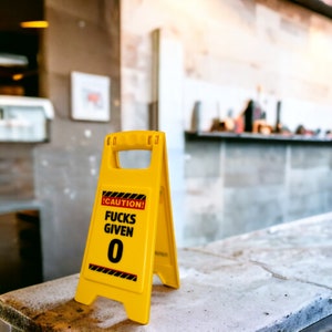 May include: A bright yellow caution sign with the text "CAUTION! FUCKS GIVEN 0" in black. The sign has a handle at the top and black and red hazard stripes. It is placed on a light-colored surface.