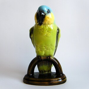 May include: A ceramic parrot figurine in shades of green, yellow, and blue. The bird is perched on a brown, arched stand with a circular base. The figurine has a glossy finish and is likely a decorative item.