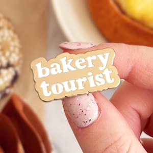 Op de afbeelding: Een beige en witte emaille pin met de tekst "bakery tourist" in een speels lettertype. De pin wordt vastgehouden in een hand met roze nagellak, met gebak op de achtergrond, waaronder een koekje en een gebakje met crèmevulling.