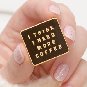Può includere: Una spilla quadrata in smalto nero con bordi dorati e la scritta "I THINK I NEED MORE COFFEE" in lettere dorate. La spilla è tenuta in mano con smalto rosa chiaro.