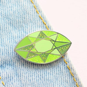 以下が含まれることがあります： 宝石の形をした緑色のグリッターエナメルピン。ピンは銀色の輪郭と薄い緑色のセンターを持っています。