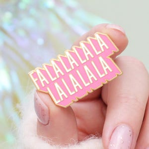 Könnte beinhalten: Ein rosa Emaille-Anstecknadel mit dem Text "FA LA LA LA LA LA LA LA" in weißen Buchstaben. Die Anstecknadel hat einen goldenen Rand und wird von einer Person mit hellrosa Nagellack gehalten.