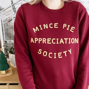 May include: A long-sleeved, crew-neck sweatshirt in a deep burgundy colour. The front features the text "MINCE PIE APPRECIATION SOCIETY" printed in a bold, light yellow, sans-serif font arranged in three centred lines.