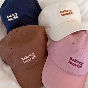 Op de afbeelding: Vier baseballpetten in marineblauw, crème, bruin en lichtroze. Elke pet is voorzien van de geborduurde tekst "bakery tourist" op het voorpaneel in een contrasterende draadkleur. De petten hebben een gestructureerde kroon en een gebogen klep.