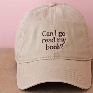 Può includere: Un cappellino da baseball beige con la scritta ricamata "Can I go read my book?" in marrone scuro. Il cappellino è realizzato in un tessuto morbido e ha una visiera curva. Lo sfondo è rosa.