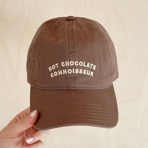 Könnte beinhalten: Braune Baseballkappe mit dem Text "HOT CHOCOLATE CONNOISSEUR" in Weiß gestickt. Die Kappe ist aus einem weichen Material gefertigt und hat einen gebogenen Schirm. Die Mütze wird von einer Person gehalten.