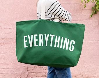 May include: A large, green tote bag with the word "EVERYTHING" printed in white. The bag has cream-colored straps and is held by a person wearing a striped sweater and jeans. The background is a pink wall.