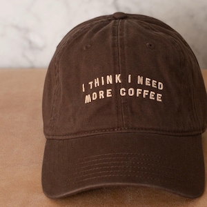 May include: A dark brown baseball cap featuring the embroidered text I THINK I NEED MORE COFFEE in a light beige colour. The cap has a structured crown, a curved brim, and visible stitching details across the panels and the front of the hat.