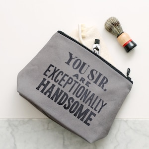 May include: Gray zippered pouch with the text "You are exceptionally handsome" printed in black. The pouch is open and contains a white washcloth, a white toothbrush, and a silver tube of toothpaste.