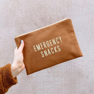 May include: A brown canvas pouch with a gold zipper and the words "EMERGENCY SNACKS" printed in gold. The pouch is rectangular and held by a person with a brown fuzzy jacket. The background is a light pink wall.