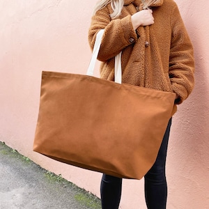 May include: A large brown canvas tote bag with white straps. The bag is being held by a person wearing a brown fluffy jacket.