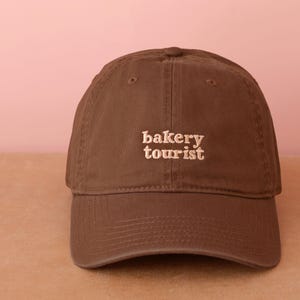 Può includere: Cappellino da baseball marrone con la scritta ricamata "bakery tourist" in beige chiaro. Il cappellino ha una visiera curva e un classico design a sei pannelli, ideale per l'uso quotidiano.