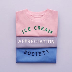 May include: Three folded t-shirts stacked on top of each other. The top shirt is pink with the text "ICE CREAM" in green letters. The middle shirt is gray with the text "APPRECIATION" in white letters. The bottom shirt is blue with the text "SOCIETY" in black letters.
