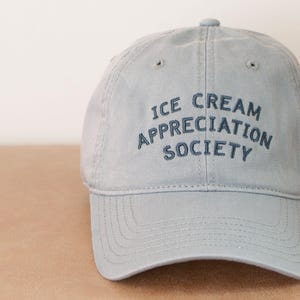 May include: A light blue baseball cap with the embroidered text "ICE CREAM APPRECIATION SOCIETY" in a darker blue. The cap has a curved brim and a fabric strap closure. The hat is made of a soft, durable material.