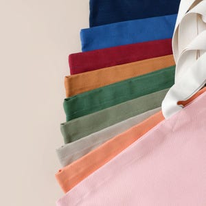 Op de afbeelding: Een stapel van tien tote bags in verschillende kleuren, waaronder roze, oranje, beige, groen en blauw. De tassen zijn gemaakt van een zacht, canvasachtig materiaal.