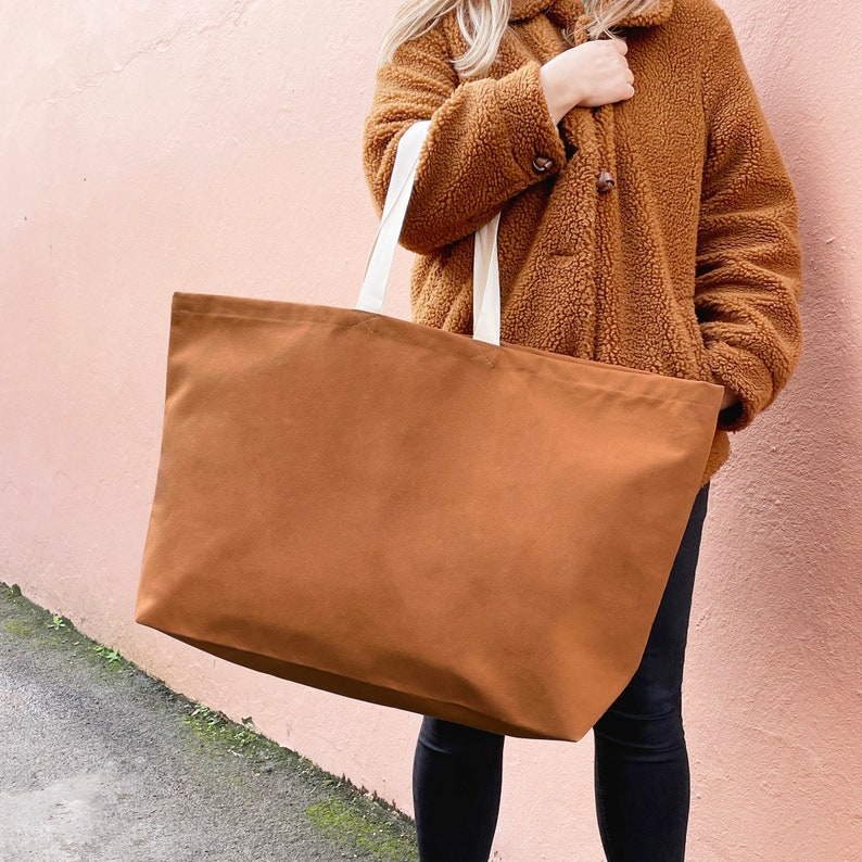 May include: A large brown canvas tote bag with white straps. The bag is being held by a person wearing a brown fluffy jacket.