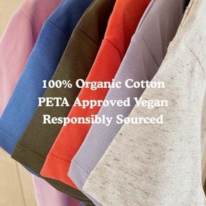 May include: A stack of six t-shirts in different colors, including pink, blue, green, red, purple, and beige. The shirts are made of 100% organic cotton and are PETA approved vegan. The text "100% Organic Cotton PETA Approved Vegan Responsibly Sourced" is printed on the shirts.